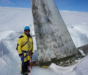 Man in harness and safety helmet stands beside aircraft tail which is rising up out of the ice