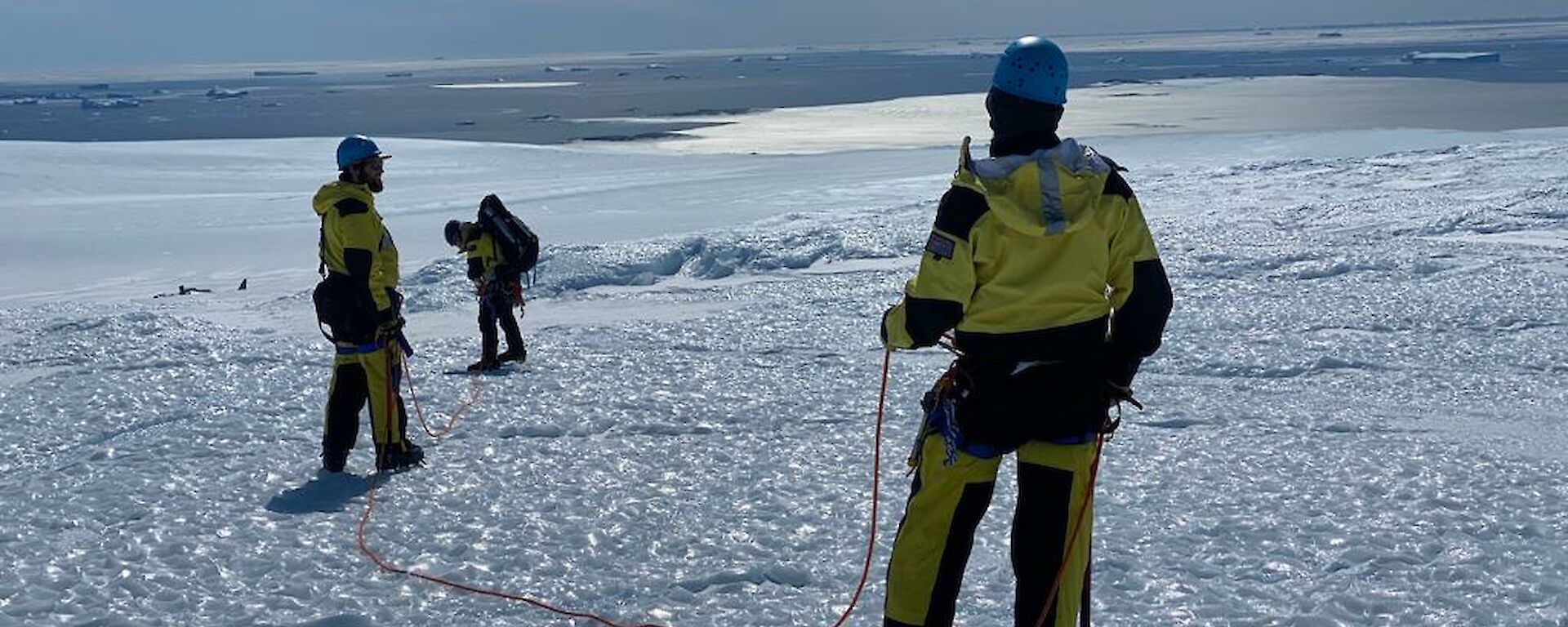 Three men wearing harness and helmets with ropes connecting them walk across icy landscape, sea in the distance