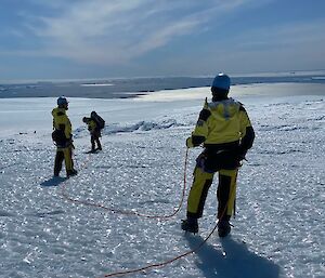 Three men wearing harness and helmets with ropes connecting them walk across icy landscape, sea in the distance