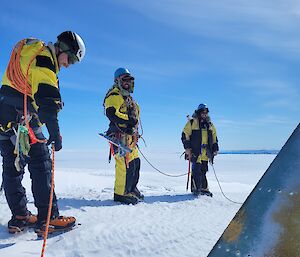 Three men in harnesses, helmets and roped together, stand on ice and look towards aircraft tail