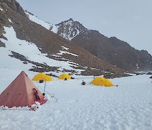 Camp site with red and yellow tents, erected on snow covered ground, with rocky mountain peak rising up in background