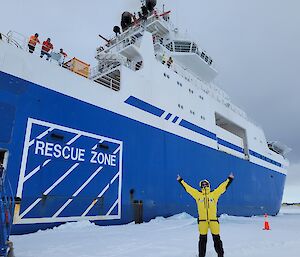 Man in AAD jacket and pants, stands on sea ice with arm raised, in front of large blue and white ship