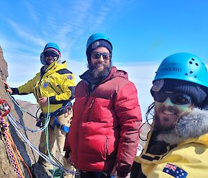 Three men in jackets, helmets and climbing harnesses smile at camera