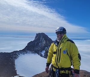 Man in AAD shell jacket, climbing harness and safety helmet smiles at camera, in the background is rocky mountain range, ice plateau and ocean in the distance