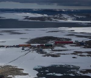 Photo from the aircraft, looking below at Casey station comprising of various red, green, yellow and blue buildings amongst the snow, with Newcomb Bay in the background.