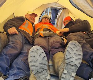 Three men in cold weather gear lie in a yellow tent