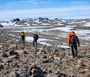 A rocky hillside with snow around and three people walking