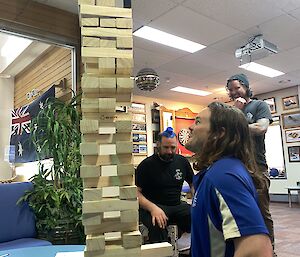 Three people stand around a large tower of wooden toy blocks