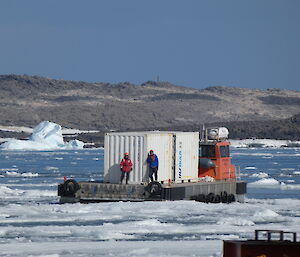 A barge with two men and a large white sea-container makes its way across sea-water filled with lots of ice-floes. land can be seen in the background.