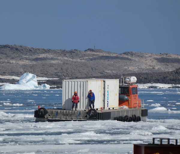 A barge with two men and a large white sea-container makes its way across sea-water filled with lots of ice-floes. land can be seen in the background.