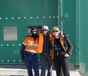 Three smiling people with their arms around one another pose for a photo outside a green building