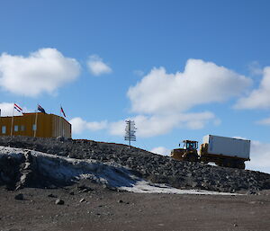 A large yellow truck with a white sea-container on the back drives up a hill towards a yellow building