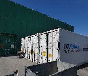 Three white sea-containers lined up side-by-side outside a large green warehouse