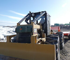 A man drives a yellow vehicle with four large tyres. It has an orange trailer attached. Sea-ice and buildings can be seen in the background.