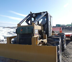 A man drives a yellow vehicle with four large tyres. It has an orange trailer attached. Sea-ice and buildings can be seen in the background.