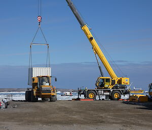 A large yellow crane loading a white sea-container onto the back of a large yellow truck. This is taking place on a wharf and the sea and a barge can be seen in the background.