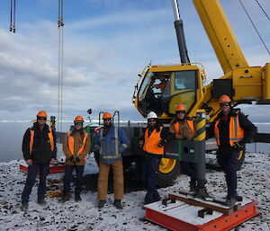 A group of six smiling men standing side by side standin in front of a yellow crane. There is a smiling man in the drivers seat of the crane. There is snow on the ground and the sea and ice-bergs can be seen in the background.