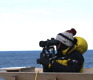 A woman dressed in a beanie and down jacket looking through binoculars while standing on a ship, with blue ocean behind.