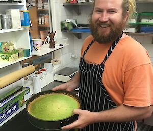 A smiling bearded man holds a green cake in a kitchen