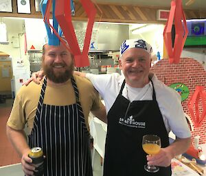Two men in aprons stand together in a kitchen with a drink in hand