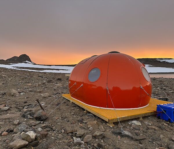 An orange melon-shaped structure pitched on a wooden platform, with a lake and snow-covered rocks in the background against a sunrise.
