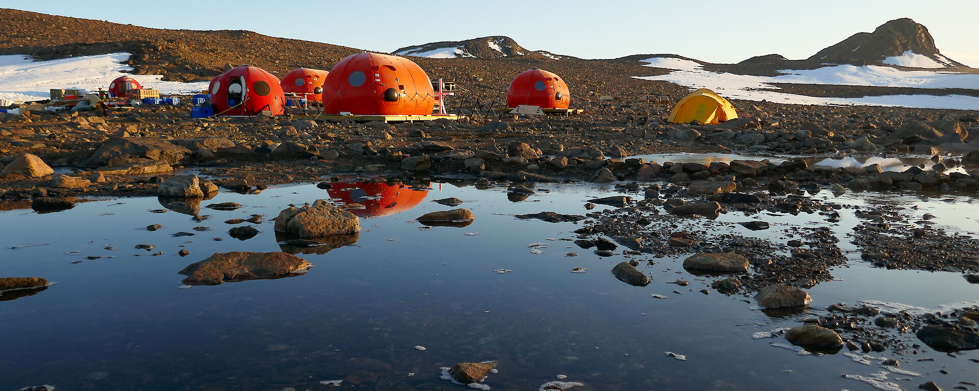 Four orange melon shaped structures behind a lake, with their reflection appearing in the lake water. Two yellow tents pitched behind them, with snow-covered rocky hills in the background.