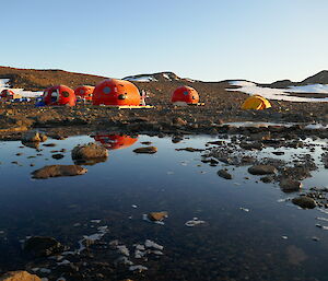 Four orange melon shaped structures behind a lake, with their reflection appearing in the lake water. Two yellow tents pitched behind them, with snow-covered rocky hills in the background.