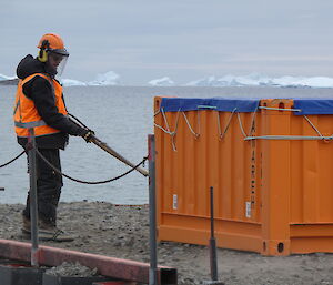 A man standing on a wharf, with an air-compressor, blows dirt off a large half-height orange sea-container