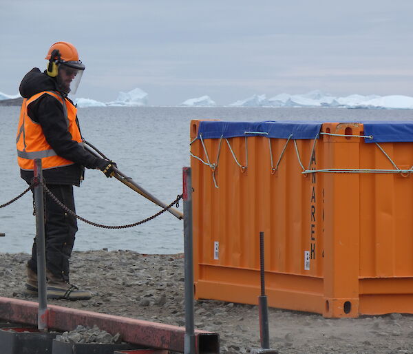 A man standing on a wharf, with an air-compressor, blows dirt off a large half-height orange sea-container