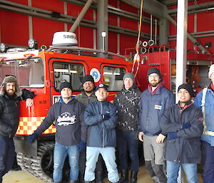 A group of 8 smiling men standing in front of a red Hägglunds