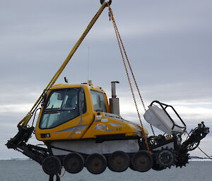 A large yellow snow groomer suspended in mid-air hanging off a crane.