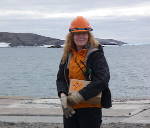 A smiling woman holding a box and wearing an orange hard-hat and a black jacket stands on a wharf with the sea in the background.