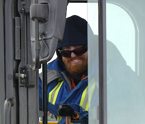 A smiling man wearing sunglasses and a hoodie sits in the cab of a vehicle.