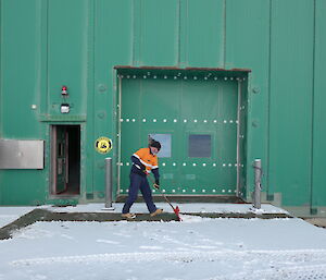 A man in high-vis clothing clearing snow from the front driveway of a large green building.