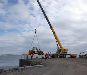 A large yellow crane lifts a smaller yellow telehandler off the ground of the wharf. Several men in high-vis clothing can be seen around the wharf and the sea is in the background.