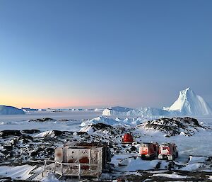 Red field huts sit on the shore with a large iceberg floating past