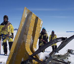 Three people stand in yellow jackets next to the broken tail wing of an old plane stuck in the snow