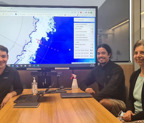Three people seated at a table with a graphic of Antarctica pictured on a large monitor behind them.