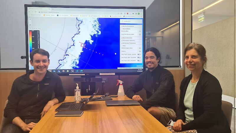 Three people seated at a table with a graphic of Antarctica pictured on a large monitor behind them.