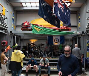 Five people in a high-ceiling shed with a skylight, setting up food and drink on a table, and musical instruments on a stage in the background. There are two Australia flags suspended from the ceiling as well as a Boxing Kangaroo flag (yellow kangaroo on a green background).