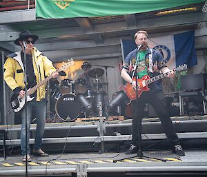 Two musicians both playing electric guitars standing on a stage with one singing into a micrphon