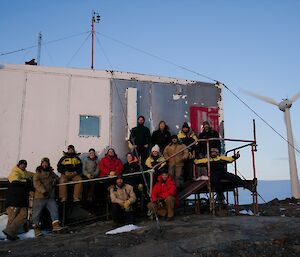 A group of people in front of a metal hut with a wind turbine behind