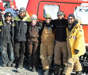Six expeditioners smile as they stand in front of a large red vehicle