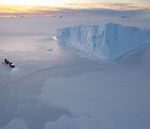 An aerial shot of a red vehicle stopped on the sea ice beside a stranded ice berg.
