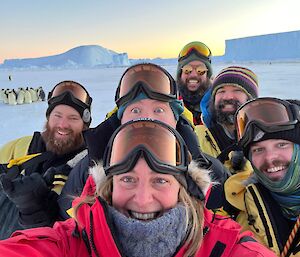 A group of people with snow goggles take a selfie in front of a penguin colony
