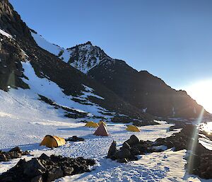 Yellow tents on the snow are dwarfed by a rocky outcrop