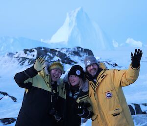 Three happy people wave a the camera in front of a large white iceberg