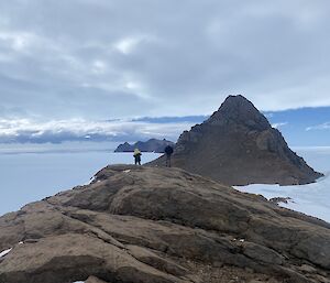 Two people stand a rocky mountain high above the sea ice