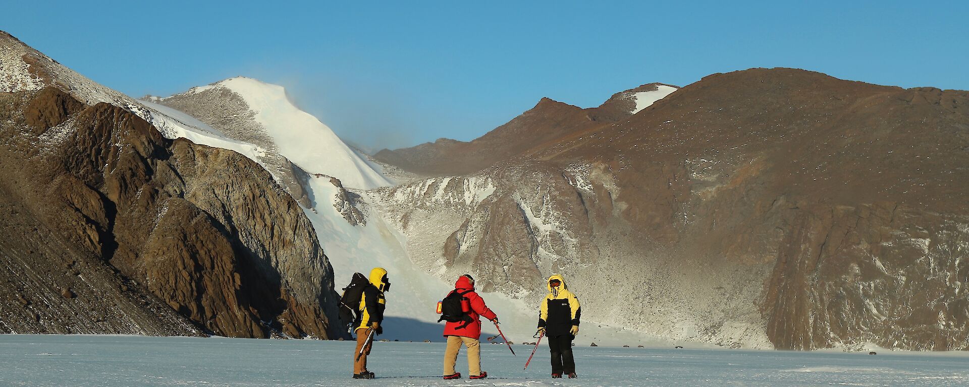 Three people stand on the snow in front of rock cliffs