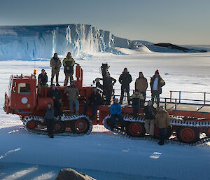 A group of people stand on a red vehicle with the sea ice and cliffs behind them